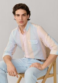 Jeune homme aux cheveux bouclés portant une chemise à rayures pastel boutonnée et un jean bleu clair, assis sur une chaise en bois devant un fond uni.
