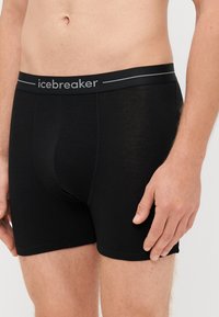 Črne bokserice iz mehkega materiala, ki imajo ožji elastični pas z napisom "icebreaker" v sivi barvi, zasnovane za udobje in prileganje.