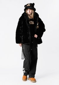 Chaqueta de piel sintética negra con mangas anchas, combinada con una camisa negra con bordado de oso, pantalones negros holgados y botas naranjas.