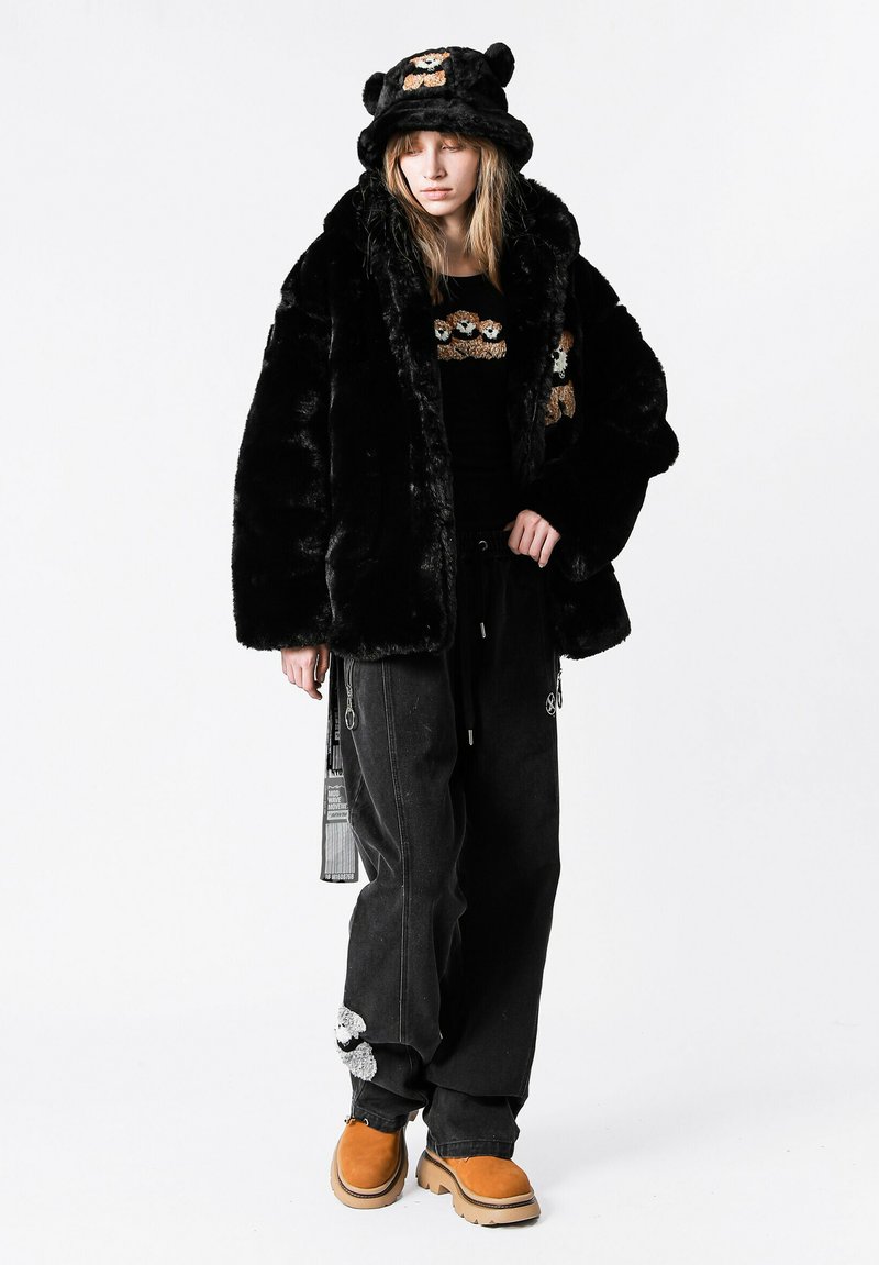 Chaqueta de piel sintética negra con mangas anchas, combinada con una camisa negra con bordado de oso, pantalones negros holgados y botas naranjas.