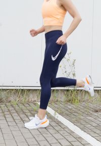 Marineblauwe sportlegging met een wit Nike-logo, gecombineerd met een perzikkleurige crop top en witte sneakers met oranje accenten, op een verharde ondergrond.