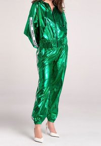 Green metallic tracksuit with a zippered jacket and elastic waistband. White branding accents on sleeves and sides. Fitted cuffs and reflective texture.