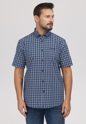 Mężczyzna z brodą w koszuli w kratę z krótkim rękawem w kolorach niebieskim i białym oraz niebieskich dżinsach, patrzący na bok.