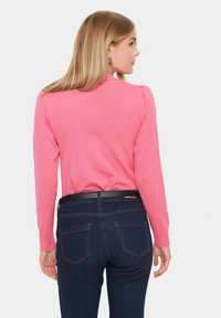 Prilagojeni pulover z dolgimi rokavi v svetlo roza pletenini, v kombinaciji s temno denim hlačami, ki vključujejo črn pas in subtilno šivanje zadnjih žepov.