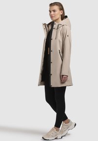 Veste imperméable beige avec capuche, double fermeture éclair et poches latérales. Associée à des leggings noirs et des baskets légères, dotées d'un design texturé.