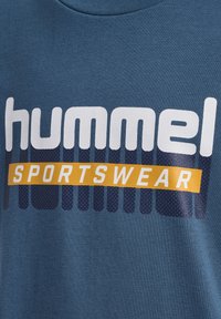 Blauwe katoen sweatshirt met gedurfde witte tekst "hummel" en "SPORTSWEAR" in oranje op een gestructureerde achtergrond.