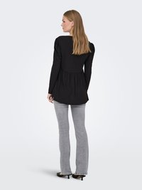 Femme aux longs cheveux blonds portant un haut noir à manches longues, un jean gris et des talons noirs, debout de dos sur fond blanc.