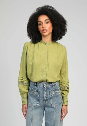 Femme aux cheveux bouclés naturels portant un chemisier vert à boutons avec des détails en dentelle et un jean bleu taille haute, debout devant un fond uni.