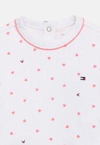 Λευκό βρεφικό φορμάκι με ροζ σχέδιο καρδιάς, μικρά λογότυπα σημαίας, ροζ δια κοσμητική μπορντούρα γύρω από το λαιμό και κούμπωμα με κουμπιά στο πίσω μέρος.