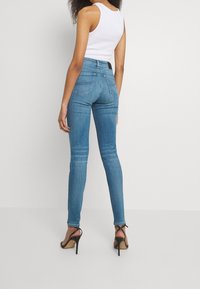 Blå skinny jeans med midja, subtil blekning och två bakfickor. Bärs med svarta klacksandaler och en vit linne.