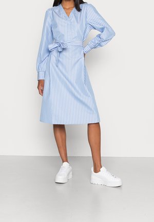 Woman wearing a blue and white vertical striped long-sleeve midi dress with a tied waist belt and white platform sneakers.