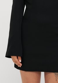 Svart minikjole med lange, utsvevende ermer; glatt tekstur; falden er rett. Hånden hviler på siden; synlig tatovering på underarmen.