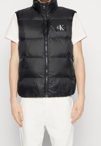 Svart pufferväst tillverkad av glänsande nylonmaterial, med hög krage, framdragkedja och sidofickor. Logopatch på bröstet.