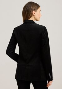 Blazer in corduroy nero con un design aderente, colletto a punta e maniche lunghe. Presenta dettagli con bottoni ai polsini e una texture liscia.