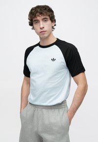 Ανοιχτό μπλε μπλουζάκι με μαύρα μανίκια ράγκλαν, με μαύρο λογότυπο Adidas στο αριστερό μέρος του στήθους. Γκρι παντελόνι φόρμας με ελαστική μέση.