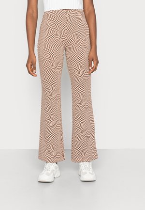 Personne portant un pantalon évasé à carreaux marron et beige taille haute et des baskets blanches épaisses, debout devant un fond uni.
