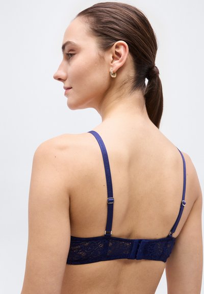 Γυναίκα με καστανά μαλλιά δεμένα πίσω, φοράει μπλε δαντελένιο σουτιέν με ρυθμιζόμενες τιράντες, εμφανίζεται από πίσω σε απλό ανοιχτό φόντο.