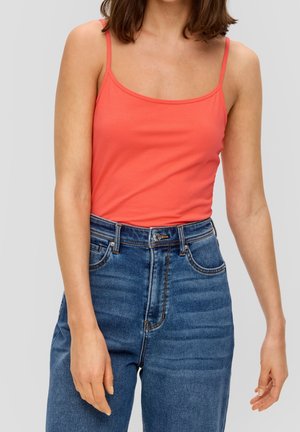 Frau trägt ein ärmelloses korallenfarbenes Oberteil und hoch taillierte blaue Jeans, steht vor einem einfarbigen hellgrauen Hintergrund.