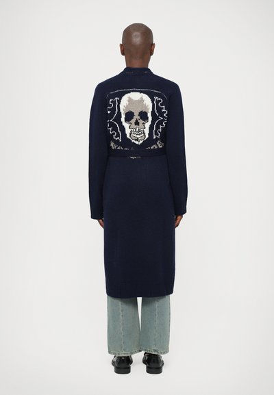 Cardigan en maille bleu marine avec un design de crâne dans le dos, manches longues et coupe décontractée, associé à un pantalon en denim clair à jambe large.