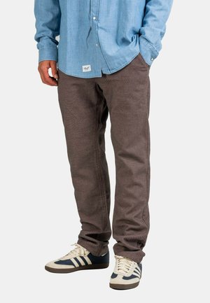 Man wearing brown textured pants, light blue buttoned shirt with label at hem, and white sneakers with navy stripes, standing with one hand in pocket.