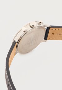 Πίσω μέρος ασημένιου ρολογιού χειρός με χαραγμένο λογότυπο και κουμπιά ρύθμισης, με μαύρο δερμάτινο λουρί με μπεζ ραφές και οπές.