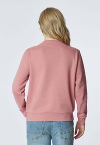 Felpa rosa chiaro con maniche lunghe e colletto a girocollo. Materiale in cotone morbido con polsini e orlo a coste, abbinata a jeans blu.