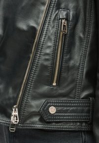 Fekete bőrdzseki közeli képe, arany cipzár részletekkel, varrott varratokkal és márkajelzéssel ellátott fém patenttal.