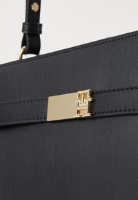 Sac à main en cuir noir avec quincaillerie dorée. Présente une texture lisse, une forme rectangulaire et un accent logo proéminent à l'avant.