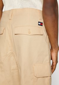 Pantalon cargo beige avec poches arrière boutonnées et poches latérales, patch logo Tommy Jeans, porté avec un haut sans manches blanc texturé.