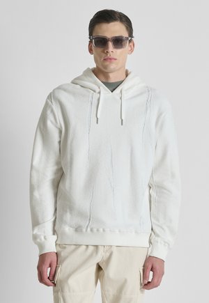 Giovane uomo che indossa occhiali da sole scuri, felpa con cappuccio bianca e testurizzata, e pantaloni cargo beige chiaro, in piedi davanti a uno sfondo chiaro e uniforme.