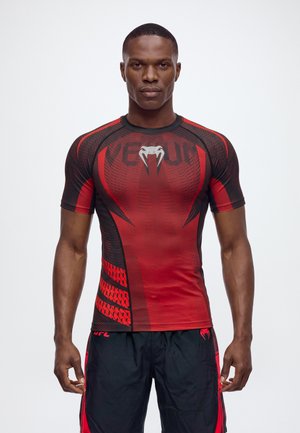 Un homme pose vêtu d’un haut de compression rouge et noir avec un logo blanc sur la poitrine et d’un short noir, sur un fond clair et uni.