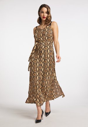 Vrouw draagt een mouwloze midi-jurk met een bruin-zwart slangenhuidprint en een strik aan de zijkant, zwarte hoge hakken en gouden sieraden, staand tegen een effen achtergrond.