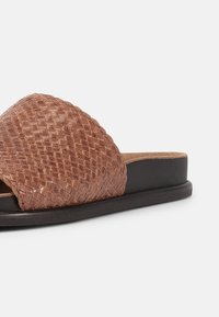 Παντόφλα τύπου σλάιντ με καφέ πλεκτό δερμάτινο πάνω μέρος και μαύρη επίπεδη σόλα, που εμφανίζεται σε προφίλ πλευράς σε λευκό φόντο.