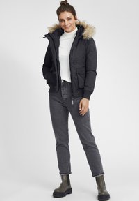 Svart parka med faux fur huva, frontdragkedja och knäppning. Buren över en vit polotröja och ihop med grå jeans och gröna ankelboots.