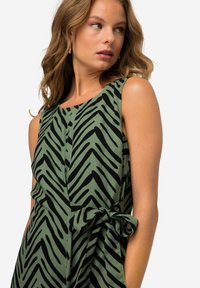 Grünes Kleid mit einem schwarzen Zickzackmuster, ärmelloses Design, gebundenem Tailleneinsatz, aus einem strukturierten Stoff gefertigt.