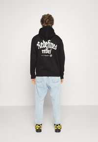 Sort hættetrøje med hvid tekst "Redefined rebel" på ryggen, kombineret med lyseblå baggy jeans og sko med gule accenter.