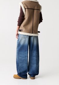 Gilet in suede marrone con bordo in shearling bianco, abbinato a jeans a gamba larga blu. Stivali chunky beige completano l'outfit.