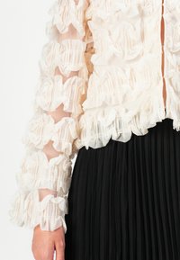 Blusa cremosa trasparente con rouches a strati, maniche lunghe e zip frontale, abbinata a una gonna nera plissettata. Il tessuto testurizzato contrasta con la superficie liscia.