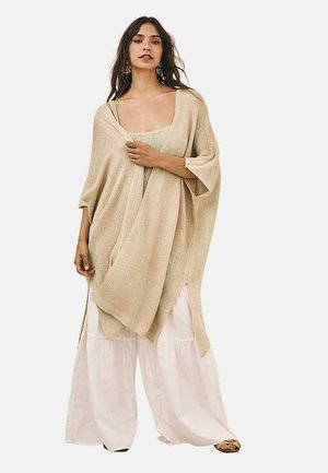 Poncho de punto beige con mangas anchas, superpuesto sobre una camiseta a juego. El modelo lleva pantalones blancos holgados y sandalias; diseño minimalista, textura ligera.