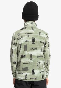 Grüne Fleecejacke mit schwarzen abstrakten Mustern, hohem Kragen und langen Ärmeln. Getragen mit einer schwarzen Strickmütze. Glatte Textur.