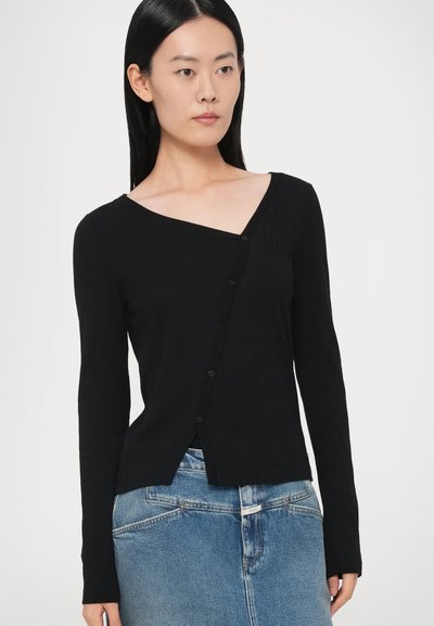 Melnas riboti garām piedurknēm džemperis ar V veida izgriezumu, asimetrisku priekšējā aizdari un pogu detaļām, kombinēts ar džinsa svārkiem.