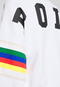 Bílá mikina s černými blokovými písmeny „AOI“. Rukáv zdobený horizontálními duhovými pruhy, které dodávají barevný akcent.
