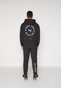 Schwarzer Hoodie und Jogginghosen mit weißem "HYROX TRAINING CLUB"-Text und Logo. Mit Kordelzug und gerippten Bündchen. Schwarze Sneakers sind sichtbar.