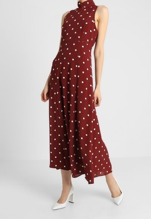 Bordeauxrotes ärmelloses Kleid mit hohem Ausschnitt, weißen Polka-Dots, fließender Silhouette und knielangem Saum. Kombiniert mit weißen High Heels.