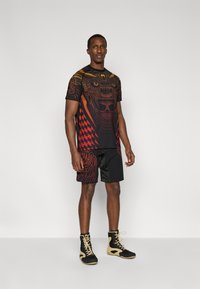 Μαύρο αθλητικό σύνολο με εφαρμοστή κοντομάνικη μπλούζα με κόκκινα και κίτρινα μοτίβα και μαύρο σορτς με αντίστοιχα γραφικά διακοσμητικά στοιχεία.