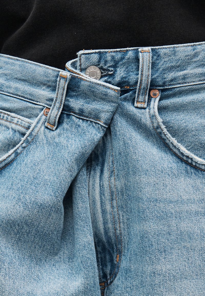 Jean en denim bleu clair avec une ceinture partiellement déboutonnée, montrant un bouton métallique et des passants de ceinture sur un fond noir.
