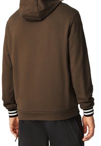 Uomo che indossa un hoodie marrone con il cappuccio sopra, pantaloni neri e polsini a righe bianche e nere visibili sui polsi.