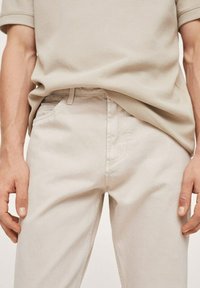 Pantalones de algodón color beige claro con un corte ajustado, que cuentan con bolsillos laterales y un tejido suave y texturizado. Se pueden ver las trabillas del cinturón en la cintura.