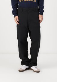 Calças cargo pretas com um ajuste solto, apresentando grandes bolsos laterais. Combinadas com um sweatshirt azul-marinho e ténis brancos com detalhes cinzas.