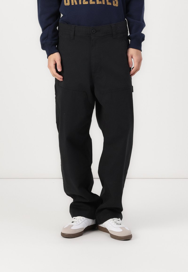 Calças cargo pretas com um ajuste solto, apresentando grandes bolsos laterais. Combinadas com um sweatshirt azul-marinho e ténis brancos com detalhes cinzas.